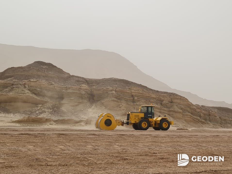 Tractor towing a non-circular impact roller across a desert site below rocky hills